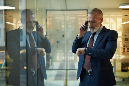 Mature Caucasian man checking time on smartwatch while talking on phone, standing in modern office with panoramic windows
