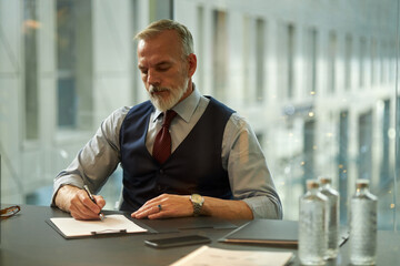 Medium shot of mature Caucasian executive sitting at table in modern board room, signing agreement...
