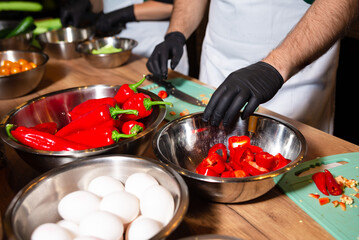 Chef slicing red peppers in kitchen prep. Gloved hands cutting fresh red bell peppers on a cutting board during kitchen prep, surrounded by other ingredients and utensils.