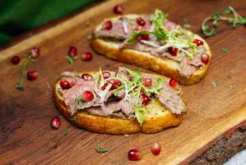 Beef bruschetta with pomegranate and greens. Toasted bread topped with sliced beef, fresh herbs, and pomegranate seeds served on rustic wooden board for gourmet presentation.