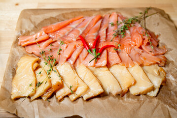 Cold smoked fish platter with herbs and chili. Assorted slices of cold smoked fish arranged on parchment paper, garnished with red chili peppers and sprigs of fresh thyme.