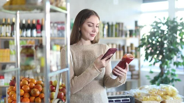 Interested young girl pondering choice of healthy and tasty ketchup, scanning barcode with smartphone to get information about product in big grocery store