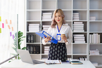 Cheerful businesswoman holding coffee cup and folders standing confidently in office environment