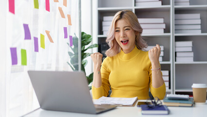 Excited businesswoman celebrating project success while working on laptop at modern office workspace