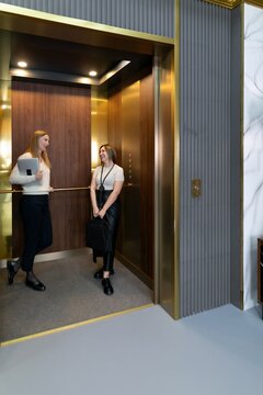 Two women engage in conversation inside a modern elevator in a stylish building during daytime hours in an urban environment
