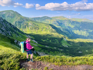 Obraz premium Woman hiker stands on mountain trail, taking in breathtaking view of rolling green hills and valleys. Backpack and sleeping pad visible as she grips trekking poles under partly cloudy sky.