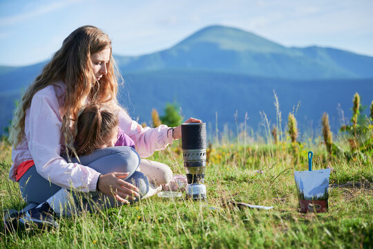 Woman prepares meal from packaged pouch next to portable camping stove. Female tourist with long blonde hair, sitting on grassy hillside with mountains in background, enjoying scenic outdoor setting.