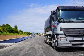Truck with bitumen spreader attachment applying hot bitumen coating on fresh asphalt with highway construction in the background
