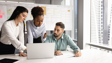 Serious young African manager presenting business plan, project strategy, talking to colleagues, showing and explaining online presentation on laptop. Diverse team working at computer together