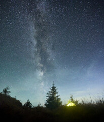 Fototapeta premium Radiant yellow tent beneath awe-inspiring, star-filled sky with Milky Way prominently stretching across. Surrounded by pine trees and silhouetted by hill, scene epitomizes tranquility of nature.