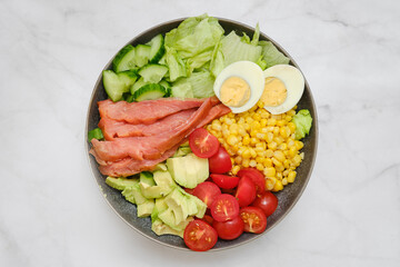 Fresh salad bowl with smoked salmon, vegetables, and boiled eggs served on a marble countertop