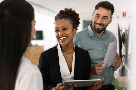 Happy diverse business partners talking at whiteboard, discussing collaboration, partnership terms, smiling, laughing, holding tablet, paper financial report, meeting for negotiation