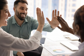 Diverse team of happy excited colleagues giving group high five over meeting table, celebrating...