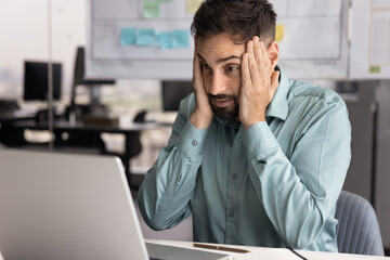 Shocked Latin businessman touching head in surprise at office workplace, looking at laptop screen, watching or reading concerning content, getting connection error, data loss message