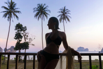 A woman stands on a balcony, silhouetted against a vibrant sunset over the ocean. Palm trees frame the tranquil scene, creating a serene atmosphere in a tropical location