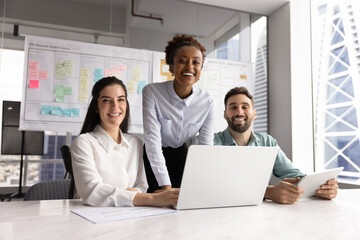 Cheerful successful diverse business team of Latin and African colleagues meeting at laptop in office workspace for teamwork, collaboration on project, looking at camera, posing for group portrait