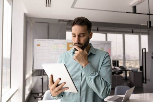 Serious thoughtful Hispanic business leader man using tablet in office workspace alone, looking ad gadget, touching chin, frowning, thinking on work challenge, problem, making decision