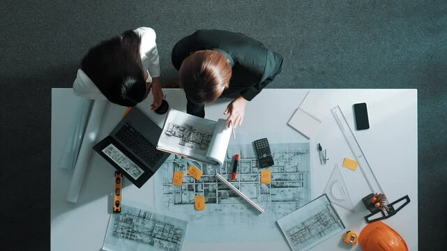 Top view of skilled project manager holding project plan and explain house design to architect engineer at meeting table with equipment, ruler, safety helmet and laptop scatter around. Alimentation.