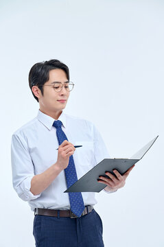 A handsome young Asian man wearing a white shirt, tie, and glasses holds a file and a pen, lost in thought.