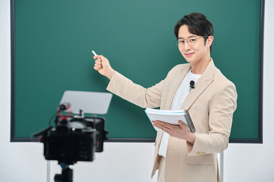 A young male instructor in a suit is holding a book and delivering an online lecture in front of the camera.