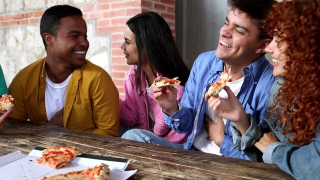 Group of multi ethnic young friends sharing pizza, laughing and enjoying casual meal together at wooden table with brick wall background