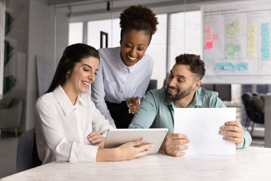 Positive multiethnic business team working on project plan together, sitting and standing at work table in office meeting space, using digital tablet for communication, smiling, laughing