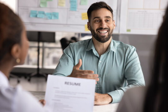 Happy confident male Latin job candidate talking to female HR managers on interview meeting, sitting at table, speaking, laughing. Recruit agents holding paper resume cropped shot
