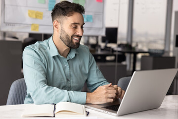 Positive Hispanic business professional man using laptop computer for job communication in office, typing, looking at screen, smiling, laughing, working on online project, smiling