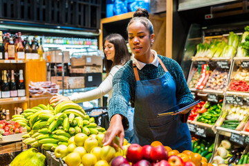 Saleswoman arranging fresh fruit in organic supermarket with customer choosing bananas