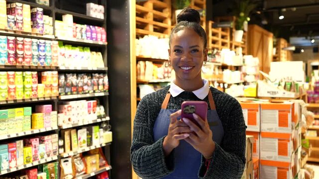 Supermarket employee using a smartphone, smiling and looking at camera, in a grocery store specialized in organic and gluten free products
