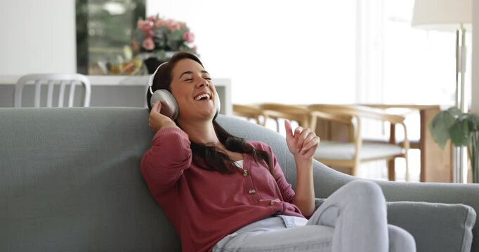 Happy inspired young Hispanic woman relaxing on home couch with closed eyes, listening o favorite music from headphones, singing song, waving hand at rhythm, smiling, laughing