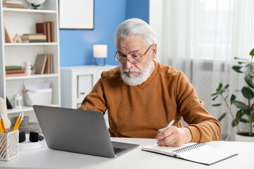 Fototapeta premium Senior man taking notes while learning online at desk indoors. Self-study