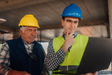 Engineers wearing safety vests and hardhats analyzing blueprints on construction site using laptop