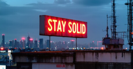 Obraz premium Glowing sign stay solid in bold red and white letters attached to rooftop ledge with city skyline behind glowing antennas and blinking red aviation lights nearby