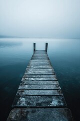 Fototapeta premium A wooden pier extends into a calm, mist-shrouded lake under an overcast sky.