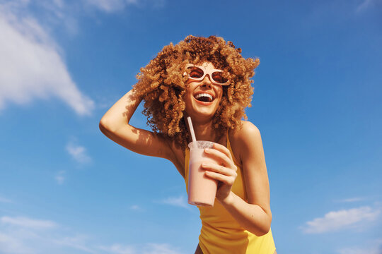 Happy young woman with curly hair enjoying a smoothie at the beach under a blue sky