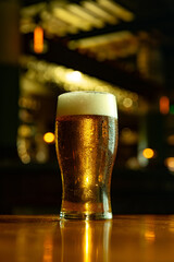 Photo of golden beer in tall pint glass with thick foam glowing under warm pub lights and blurred bottles on bar in background. Concept of alcohol, festival, beer, culture, pub, bar.
