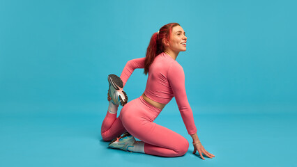 Flexible young woman in pink outfit stretching quadriceps in kneeling pose, smiling and relaxed, showing warm-up routine. Concept of healthy spine, mobility, active lifestyle and workout habits.