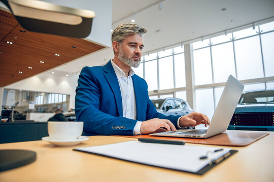 Bearded salesman in suit working on his laptop at new cars showroom