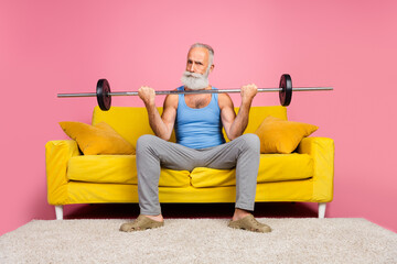 Senior man lifting weights on a couch in a pink room showcasing leisure and strength