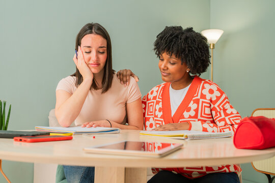 Two diverse young women studying together in a cozy indoor setting while one offers emotional support to the other during a difficult academic challenge at a light-filled table