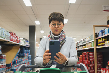 Focused senior customer using a smartphone while pushing a shopping cart in a supermarket aisle, checking her shopping list or comparing prices