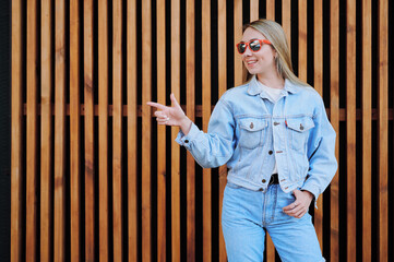 Obraz premium Smiling young woman in denim jacket and jeans pointing upwards with one hand while standing confidently against wooden slatted wall outdoors