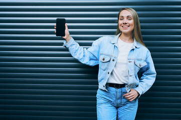 Playful young woman in denim jacket raising smartphone in one hand while smiling confidently...