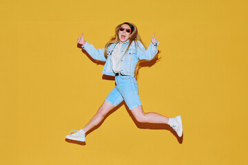 Obraz premium Dynamic woman in denim jacket and shorts jumping mid-air against bright yellow wall, showing thumbs up with both hands and smiling, capturing joyful youthful energy