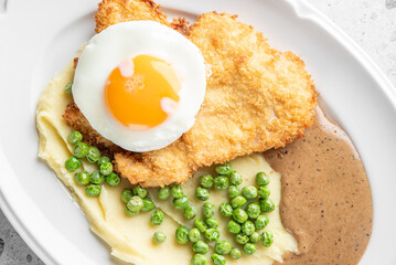 A plate of crispy fried chicken topped with a sunny-side-up egg, served with creamy mashed potatoes, green peas, and a savory brown sauce.