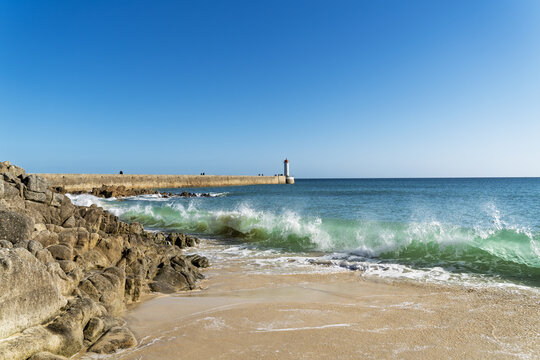 Vague turquoise sur le sable dor&eacute; face au phare et au m&ocirc;le du Raoulic &agrave; Audierne. Sous un ciel bleu, la mer scintille tandis que des promeneurs marchent sur la jet&eacute;e.