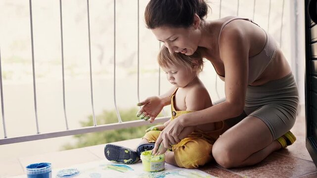 Young mother with her little boy mixing colors at hands yo leave beautiful handprints on the paper. Mother and child concept. Slowmotion. Mom amd child spend time together. Family, parenthood concept