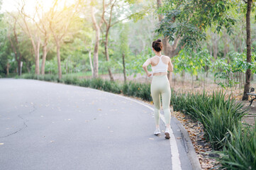 young Asian woman jogging on peaceful park pathway surrounded by lush greenery and sunlight,...