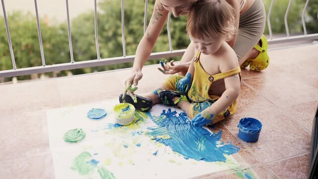 Young mother with her little boy mixing colors at hands yo leave beautiful handprints on the paper. Mother and child concept. Slowmotion. Mom amd child spend time together. Family, parenthood concept